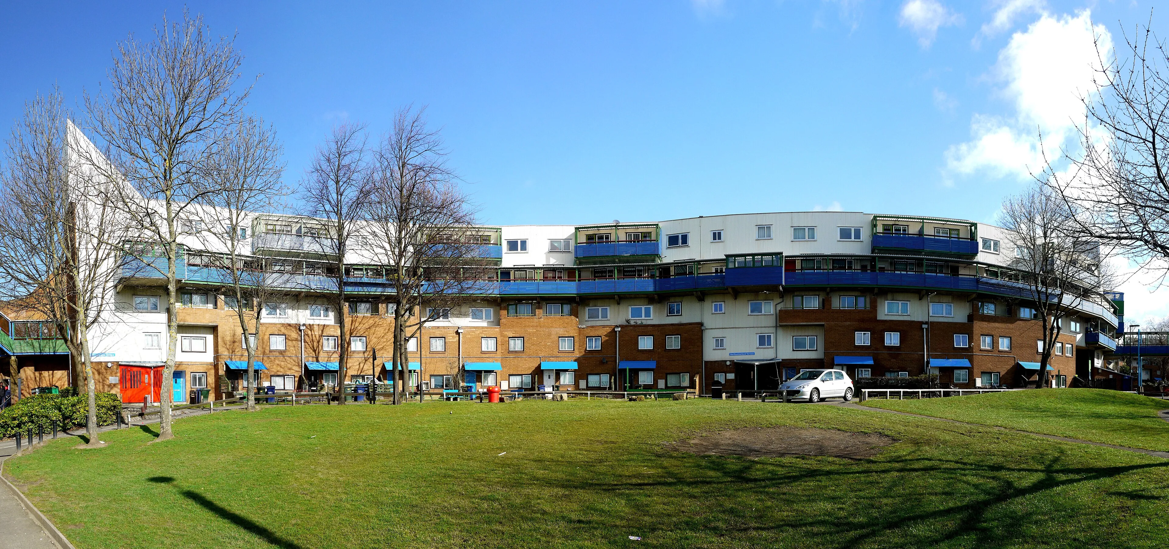 Byker Wall’s curving block of flats with brick lower floors, white upper levels and blue balconies, seen across a grassy open space under a blue sky. By Andrew Curtis, CC BY-SA 2.0, https://commons.wikimedia.org/w/index.php?curid=14566783