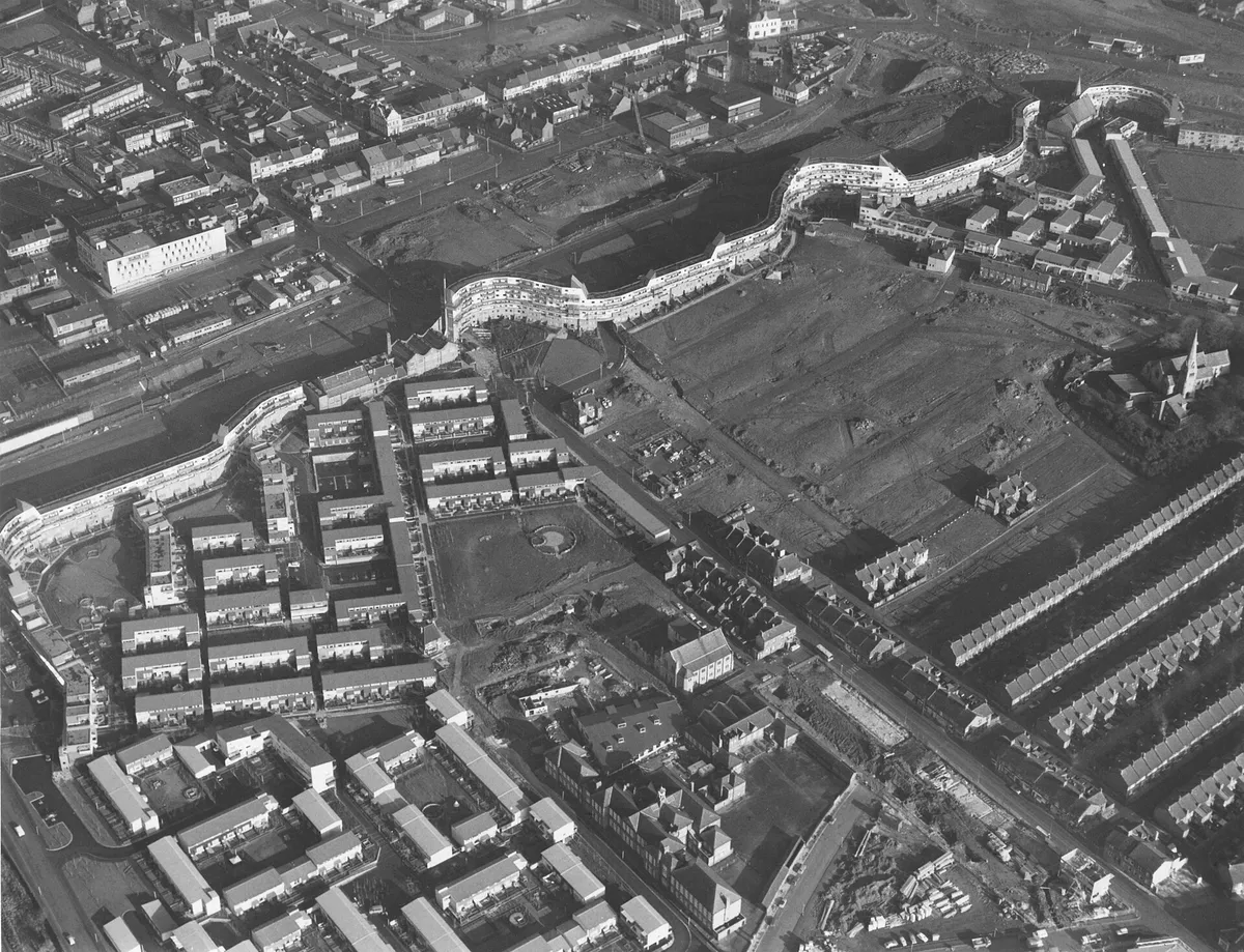 Black and white aerial photograph of Byker Wall partly built, a long curving block of flats winding across the site, with new low-rise housing in front and cleared land and terraced streets beyond.